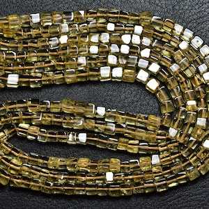 Può includere: Una collana di perle di citrino giallo chiaro, ogni perla è sfaccettata e di forma quadrata. Le perle sono infilate su un filo sottile e sono disposte in un motivo sciolto e fluido.