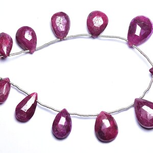 May include: A strand of ruby gemstone beads, each bead is a teardrop shape and has a faceted finish. The beads are strung on a silver wire and have pink tassels on the ends.