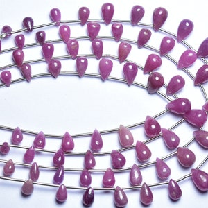 May include: A strand of pink gemstone beads in a teardrop shape. The beads are strung on a silver wire and are arranged in a circular pattern.