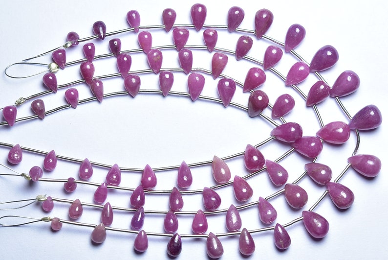 May include: A strand of pink gemstone beads in a teardrop shape. The beads are strung on a silver wire and are arranged in a circular pattern.