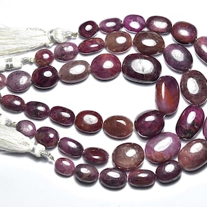 May include: Strands of polished, oval-shaped ruby gemstones in varying shades of red and purple. The gemstones are strung together, with white tassel accents at the ends. The beads are approximately 1 cm in length.