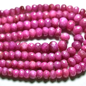 Könnte beinhalten: Eine Strähne rosafarbener Rubinperlen, facettiert und rund, mit polierter Oberfläche. Die Perlen sind auf einem Draht aufgefädelt und bereit für die Schmuckherstellung.