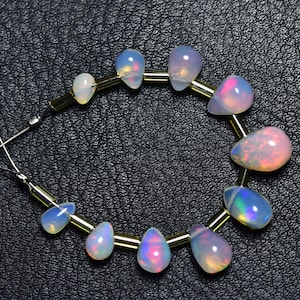 Puede incluir: Una hebra de cuentas de ópalo en forma de lágrima iridiscente enhebradas en un alambre dorado. Las cuentas son de varios tamaños y tienen un brillo arcoíris.