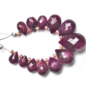 Può includere: Una collana di perline di rubino rosso scuro sfaccettate. Le gemme a forma di goccia sono infilate con piccole perline color oro e un filo sottile marrone-rosato. Le perline sono disposte in un motivo circolare su sfondo bianco.