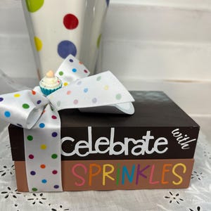 May include: A brown wooden block set with the words "celebrate with SPRINKLES" in white and rainbow colors. The set is tied with a white ribbon with colorful polka dots and a small cupcake decoration.