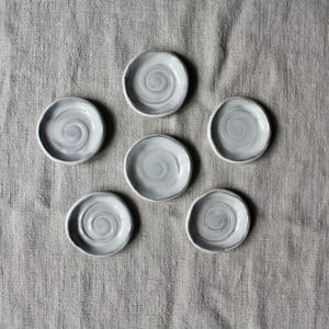 Puede incluir: Seis platos pequeños y redondos de cerámica dispuestos en un patrón circular. Cada plato presenta un esmalte gris claro con un sutil diseño de espiral gris más oscuro centrado en el medio. Los bordes son ligeramente irregulares.