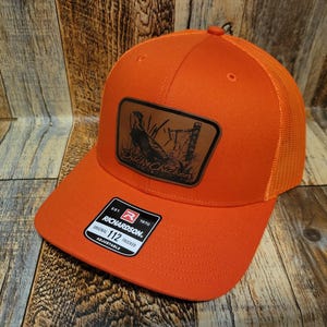 May include: Orange trucker hat with a brown leather patch featuring a pheasant and the text "Dick'n Chicken". The hat has a mesh back and a Richardson 112 adjustable strap.