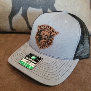 Puede incluir: Una gorra de camionero gris y negra con un parche de cuero marrón que dice "Second Amendment" y presenta una calavera con rifles cruzados. El parche también dice "Don't Tread on Liberty".