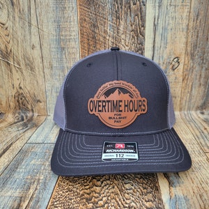 Könnte beinhalten: Eine schwarze und graue Trucker-Kappe mit einem braunen Leder-Patch, auf dem "Been Sellin' My Soul Workin' all day" und "OVERTIME HOURS FOR BULLSHIT PAY" steht.
