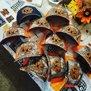 May include: A collection of camouflage and black baseball caps with a brown leather patch featuring a deer and the text "Bucks & Tracks Taxidermy".