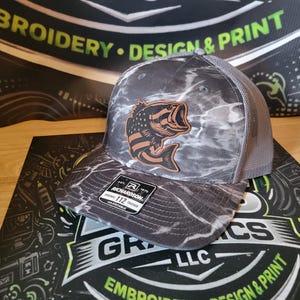 May include: A grey and white marble-patterned trucker hat featuring a brown leather patch with a black fish graphic. The hat has a mesh back, a flat brim, and a black sticker on the brim that reads Richardson 112 Original Trucker Adjustable.