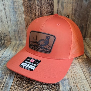 May include: Orange and black mesh trucker hat with a brown leather patch featuring a pheasant and the text "Ditch Chicken". The hat is adjustable and has a Richardson 112 label.
