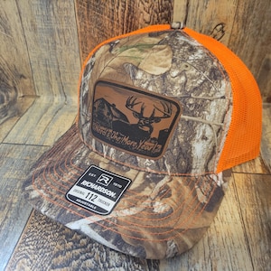 Op de afbeelding: Een camouflage en oranje trucker cap met een bruin leren patch waarop "Need One More Year" staat met een hert silhouet. De pet is gemaakt door Richardson 112.