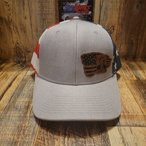 May include: A gray trucker hat with a red, white, and blue American flag design on the side. The hat has a brown leather patch with an American flag and the text "Trump 2020" on the front.