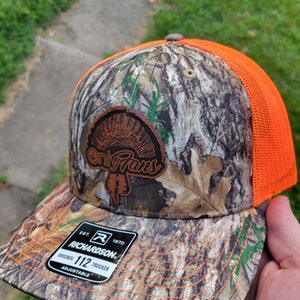 May include: A camouflage trucker hat with an orange mesh back. The hat features a brown leather patch with a turkey design and the text "Only Fans". The hat is adjustable and has a Richardson label with the text "Est. 1970 Original 112 Trucker Adjustable".