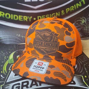 Puede incluir: Una gorra tipo trucker de camuflaje naranja y marrón con un parche de cuero sintético marrón en la parte frontal que muestra un faisán y el texto DITCH CHICKEN. Una pegatina blanca en la visera muestra el texto 112PFP PRINTED 5 PANEL OSFM.