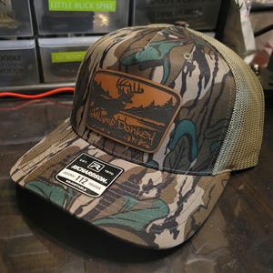 May include: A camouflage trucker hat with a brown leather patch that says "Swamp Donkey" and a buck silhouette. The hat is adjustable and has a mesh back.