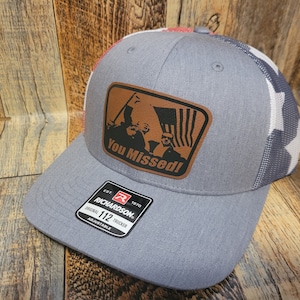 May include: A gray and white trucker hat with a brown leather patch that says "You Missed!" The patch features a black silhouette of two people holding an American flag. The hat is adjustable and has a red, white, and blue logo that says "Richardson 112 Original Trucker".