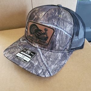 May include: A camouflage trucker hat with a brown leather patch featuring a turkey illustration and the text "Pure Grit". The hat is black mesh on the back and has a black adjustable strap with the text "Richardson 112 Trucker Adjustable".