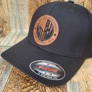 May include: Black baseball cap with a brown leather patch featuring a Spartan helmet and the text "We Are Not Descended From Fearful Men" with a small American flag. The patch also has a star on the bottom right corner. The cap has a Flexfit label on the back.