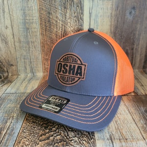 Könnte beinhalten: Eine graue und orangefarbene Trucker-Kappe mit einem braunen Leder-Patch, auf dem "Habitual OSHA Violator" steht.