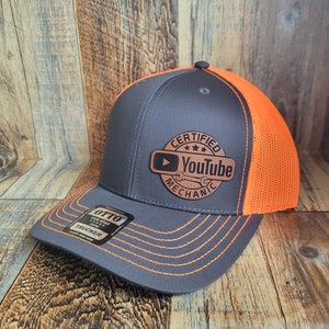 May include: A gray and orange trucker hat with a brown leather patch that says "Certified YouTube Mechanic" with a wrench and play button icon.