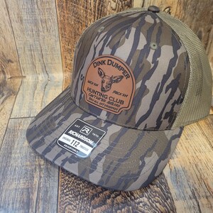 May include: A brown and green camouflage trucker hat with a brown leather patch that says "Dink Dumper" and "Hunting Club" with a deer illustration. The hat is made by Richardson and is adjustable.