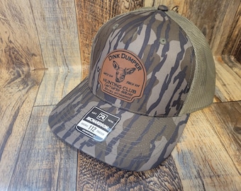 May include: A brown and green camouflage trucker hat with a brown leather patch that says "Dink Dumper" and "Hunting Club" with a deer illustration. The hat is made by Richardson and is adjustable.