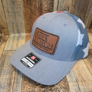 May include: A grey and blue trucker hat with a brown leather patch that says "HAWK TUAH'24 - Spit On That Thang" and a red, white and blue mesh back. The hat is made by Richardson and is size 112.