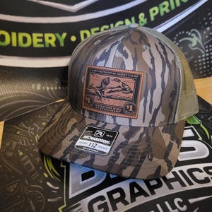May include: Camouflage trucker hat with a brown and green pattern. Features a leather patch with a vintage stamp design and the text "DEPARTMENT OF AGRICULTURE". The hat has a mesh back and a curved brim. The brand "RICHARDSON" is visible.