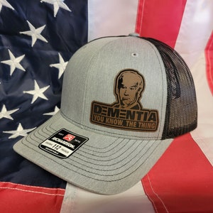 Peut inclure: Un chapeau de camionneur gris et noir avec un patch en cuir marron qui dit "DEMENTIA YOU KNOW, THE THING". Le patch représente une caricature d'une personne.