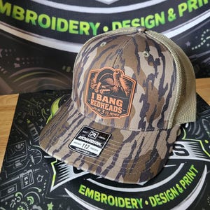 May include: Camouflage trucker hat with a brown and orange patch that reads "I BANG REDHEADS at 3 1/2 inches". The hat has a mesh back and a curved brim. The brand is Richardson 112.