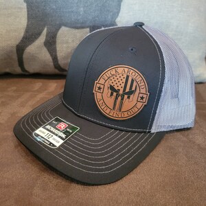 May include: A black and gray trucker hat with a brown leather patch that says "F*CK AROUND AND FIND OUT" with a skull and crossbones and an American flag. The hat is made by Richardson.