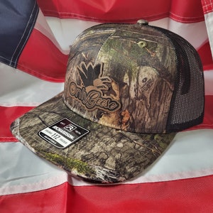 May include: A camouflage trucker hat with a brown leather patch that says "Only Goose" and a black mesh back. The hat is sitting on an American flag.