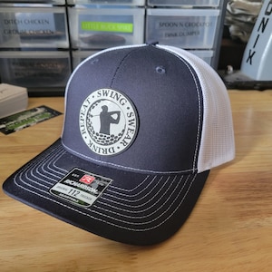 Pode incluir: Boné de caminhoneiro azul marinho e branco com aba curva. O boné apresenta uma parte traseira de malha branca e um patch circular com a silhueta de um jogador de golfe e as palavras "Repeat, Swing, Swear, Drink". O boné é ajustável.