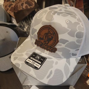May include: White camouflage trucker hat with a brown leather patch that says "Vote Smoker". The patch features a silhouette of a deer in a crosshair. The hat is adjustable and has a mesh back.