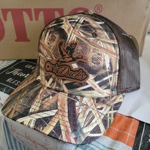 May include: A brown and tan camouflage baseball cap with a brown mesh back. The cap features a brown leather patch with the text "Only Ducks" in white lettering and a silhouette of a duck.