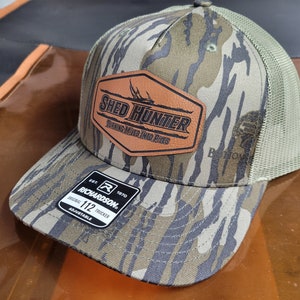 May include: A camouflage trucker hat with a brown leather patch that says "Shed Hunter Turning Hides Into Pipes". The hat is made by Richardson 112 Trucker.