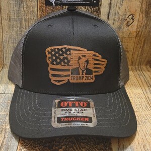 May include: A black and gray trucker hat with a brown leather patch featuring an American flag and a portrait of Donald Trump with the text "TRUMP 2024". The hat has a mesh back and a classic fit.