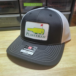 Puede incluir: Gorra de camionero negra y blanca con un parche con una botella amarilla y un diseño de campo de golf, con la palabra "PLASTERED". La gorra tiene una visera curva y una parte trasera de malla. La gorra es ajustable.