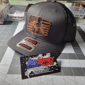 May include: A black trucker hat with a brown leather patch featuring a welder in front of an American flag. The hat has a mesh back and a label that reads "OTTO FIVE STAR TRUCKER".