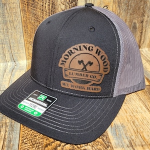 May include: A black and gray trucker hat with a brown leather patch that says "Morning Wood Lumber Co. We Work Hard".