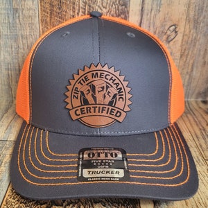 Könnte beinhalten: Ein schwarzer und orangefarbener Trucker-Hut mit einem braunen Leder-Patch, auf dem "Zip Tie Mechanic Certified" steht. Der Hut hat ein Netzrücken und einen klassischen Netzrücken.