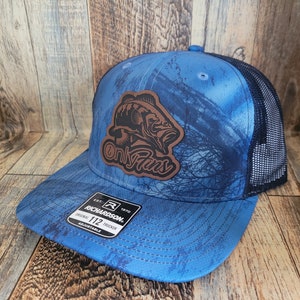 Op de afbeelding: Een blauwe en zwarte camouflage trucker cap met een bruin leren patch met een baars en de tekst "Only Fins". De pet is verstelbaar en heeft een Richardson 112 label.