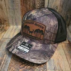 Pode incluir: Boné de caminhoneiro camuflado com costas de malha preta e um patch de couro marrom. O patch apresenta o texto "FREEZER QUEEN" acima de um gráfico de veado e árvores, com a frase "SHE BLOWS SHE GOES" abaixo. O boné é ajustável.