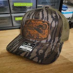 Op de afbeelding: Truckerpet met camouflagepatroon en een bruin en oranje patch met de tekst "Limb Chicken". De pet heeft een gebogen klep en een mesh achterkant. De pet is verstelbaar en heeft een Richardson 112 Trucker-label.