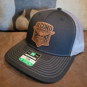 May include: A black and gray trucker hat with a brown leather patch that says "Second Amendment Freedom & Liberty" with a skull and crossbones design. The hat is made by Richardson 112RE.