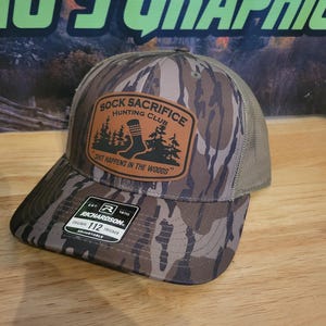 Puede incluir: Gorra de camionero de camuflaje con un patrón marrón y beige. El sombrero presenta un parche con el texto "Sock Sacrifice Hunting Club" y la frase "Shit Happens in the Woods". La gorra es ajustable.