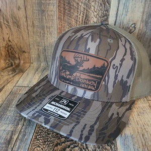 Pode incluir: Um chapéu de caminhoneiro de camuflagem marrom e bege com um remendo de couro marrom que apresenta um veado e o texto "Swamp Donkey". O chapéu é feito pela Richardson e tem tamanho 112.