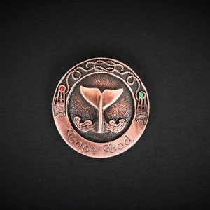May include: A copper-colored round metal pin with a whale tail design, rope border, and the words "Cape Cod" inscribed around the edge. The pin has red and green gemstones on either side of the whale tail.